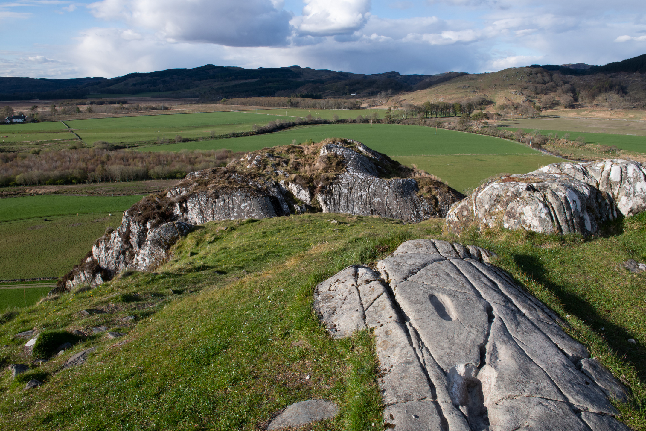 A carved footprint and other shallow rock markings on a flat stone at the top of Dunadd Hill, with fields and hills in the distance.