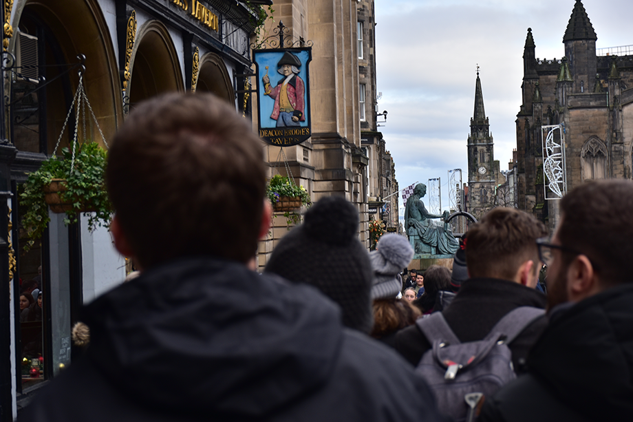 A crowd of people walking down a street next to a pub with a green statue in the distance.