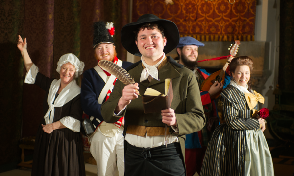 A group of five performers in 18th century Scottish costumes smile at the camera whilst standing in an ornate palace room. In the centre a man is dressed as Robert Burns, holding a leather notebook and feather quill.