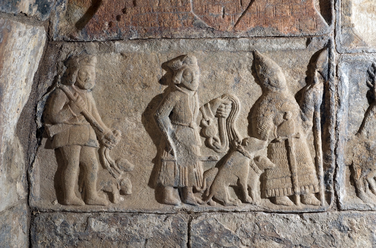 Stone carving of several figures and a dogs, depicting a medieval scene with people in traditional clothing.