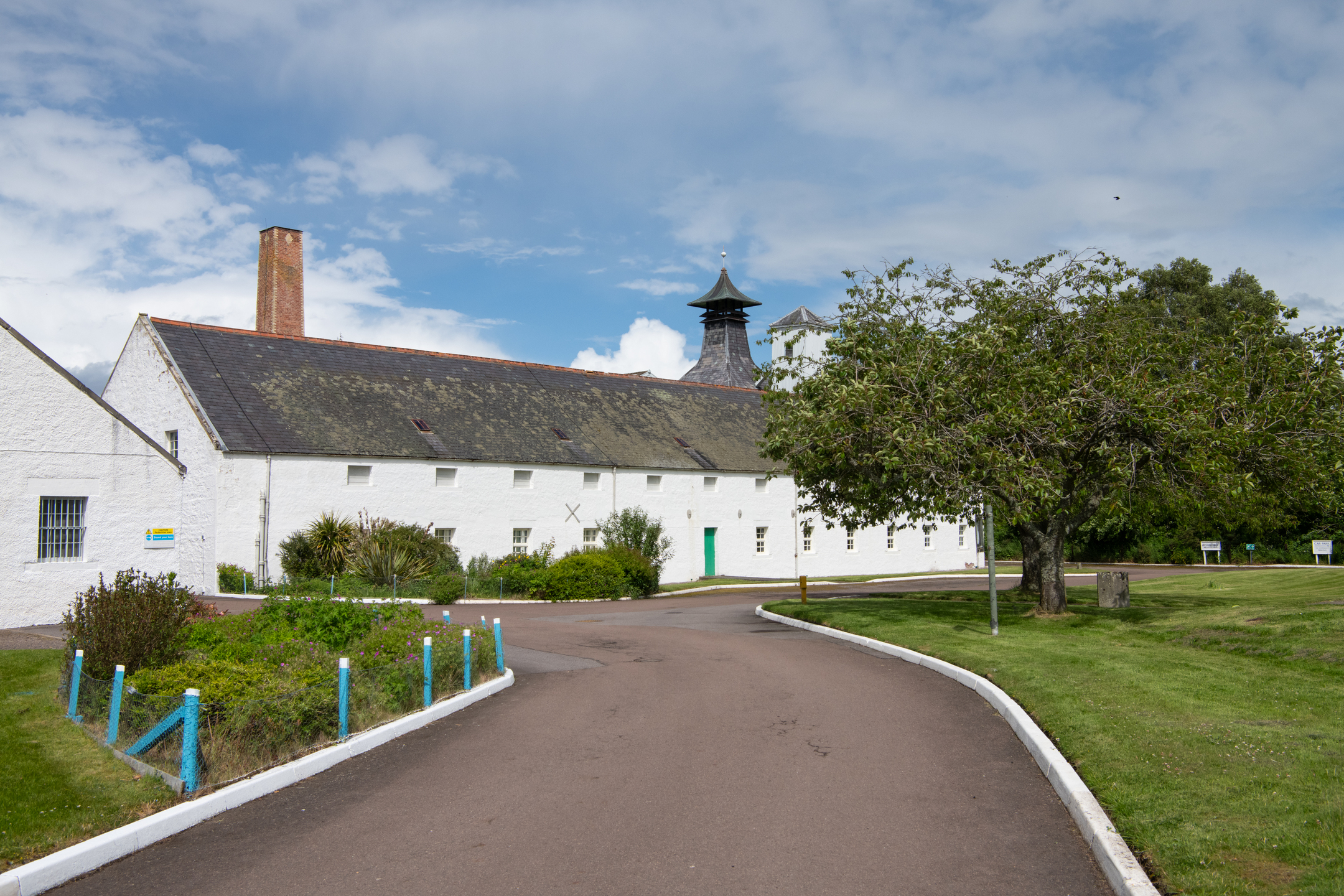 Approach road and gardens at Dallas Dhu distillery, leading past whitewashed warehouse buildings.