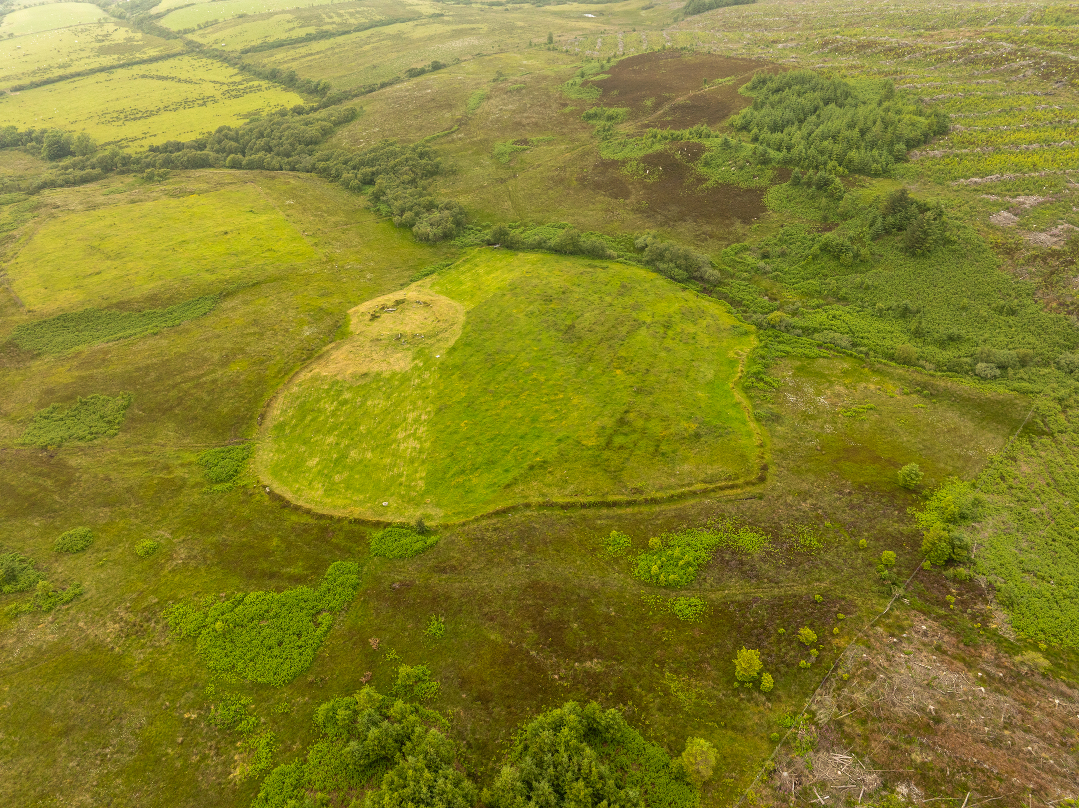 Aerial view of a large oval-shaped earthwork surrounded by moorland and patches of green vegetation, with hills visible in the distance.