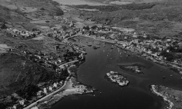  An oblique aerial image in black and white of Tarbert’s historic harbour. Several ships and boats are in the harbour and there are two islets in the middle.