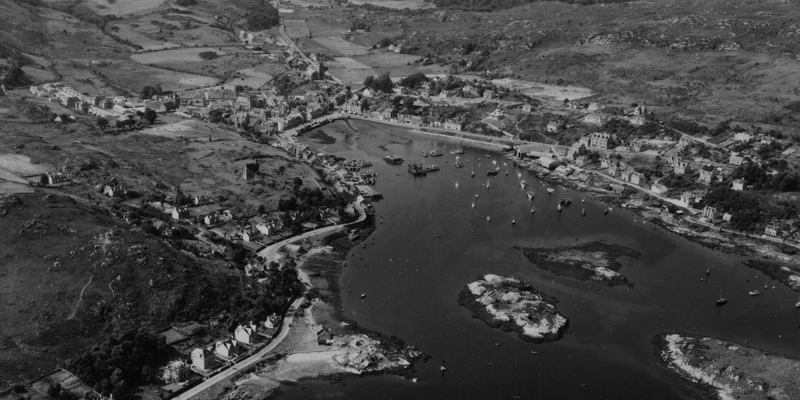  An oblique aerial image in black and white of Tarbert’s historic harbour. Several ships and boats are in the harbour and there are two islets in the middle.
