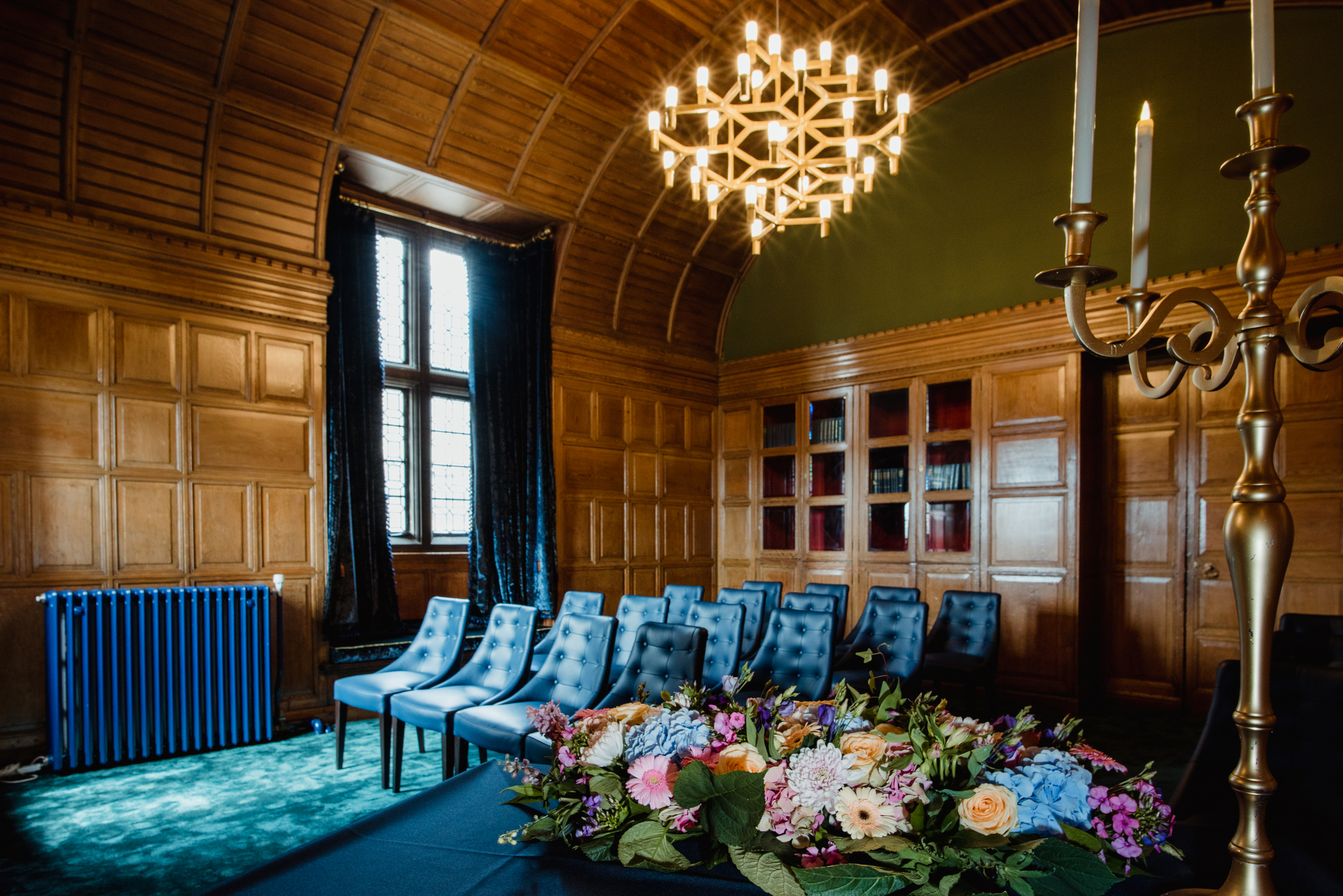 Blue chair arranged for guests to watch a ceremony. The room is covered in wood paneling and a golden chandelier lights up the room. 