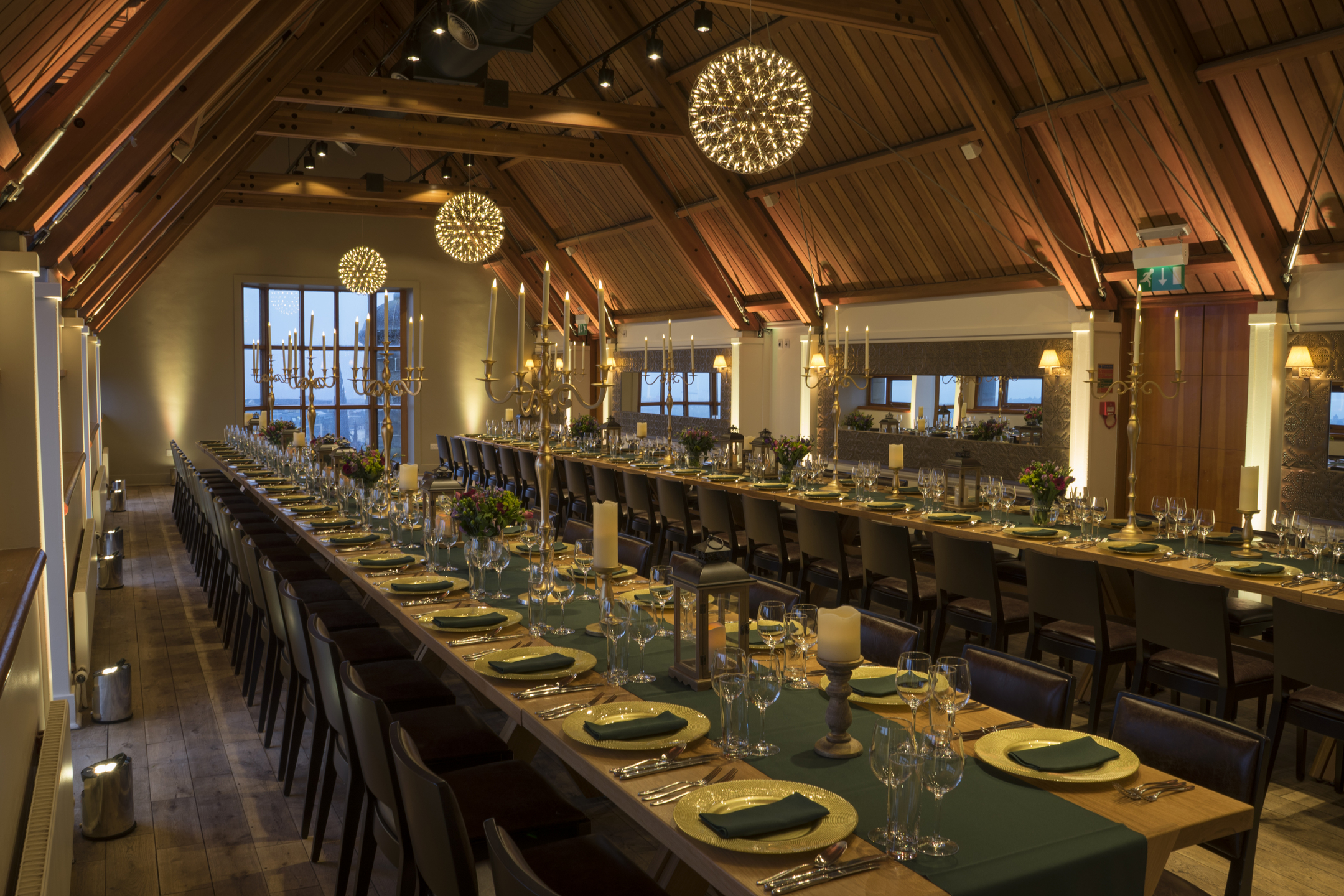 Tow very long tables are set ready for dinner guests to arrive. The long rectangular room has a high a-frame wooden ceiling and windows on every wall.