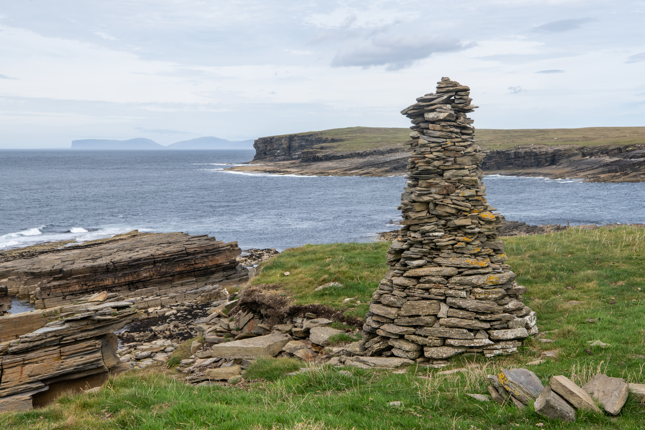 A rugged coastal landscape with a tall, conical cairn made of stacked flat stones standing on a grassy headland. To the left, sharply layered rock formations descend toward the sea, where waves break against the cliffs. The coastline curves into the distance, with low cliffs and open moorland under a pale, cloud‑covered sky. Faint outlines of distant islands appear on the horizon.