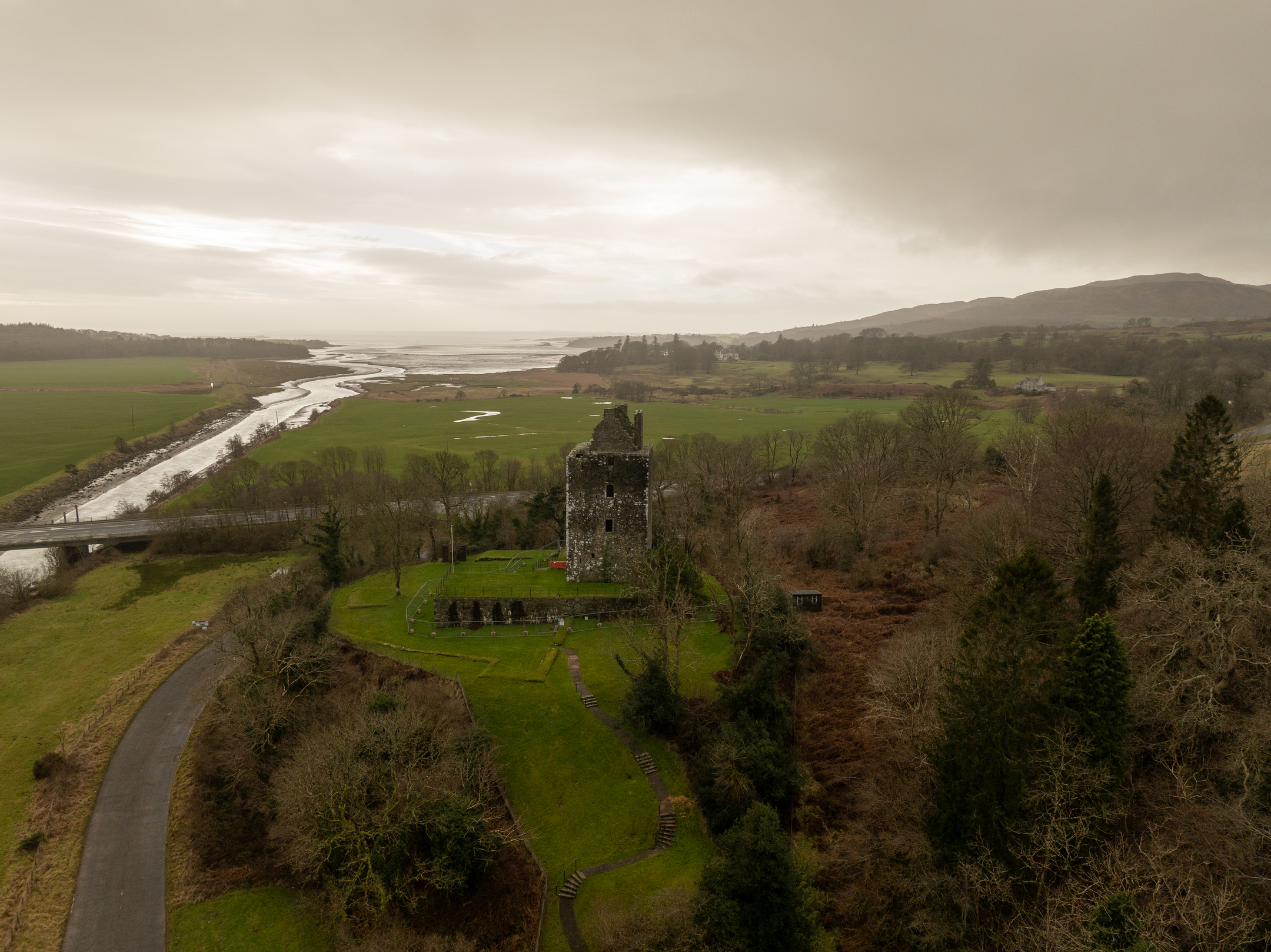 Aerial view of the stone tower overlooking expansive green fields and a meandering river flowing toward the sea under an overcast sky.
