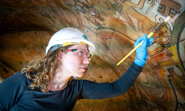 A young woman, wearing protective wear and sterile gloves, is reaching up with a paintbrush to paint an ornately decorated ceiling.