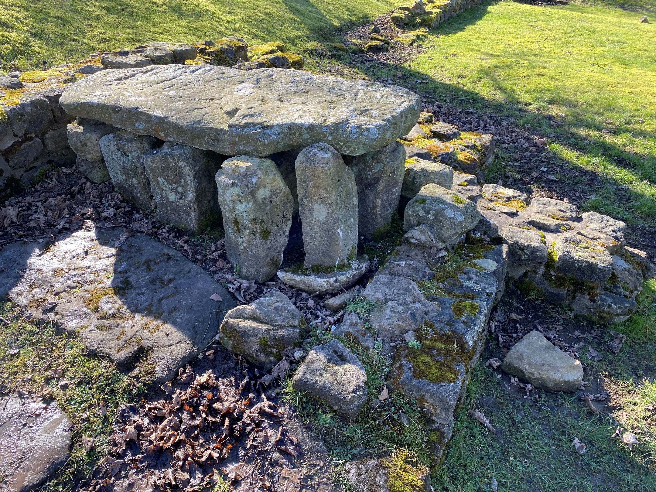 Remains of a Roman bathhouse feature at Bar Hill Fort, consisting of a large flat stone slab supported by upright stones, forming a table-like structure. Surrounding it are moss-covered stones arranged in a rectangular layout, with fallen leaves scattered on the ground. The area is grassy and slightly sloped, with sunlight casting shadows.