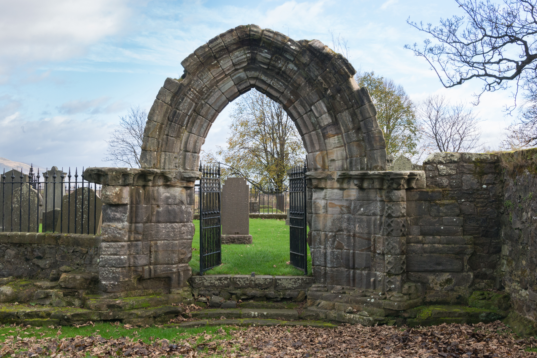 A medieval arched stone doorway leading into the Cambuskenneth Abbey burial ground. The arch is constructed of weathered stone blocks with layered mouldings and sits between sections of ruined wall. Behind the doorway, iron gates and gravestones are visible, with trees and grass beyond.