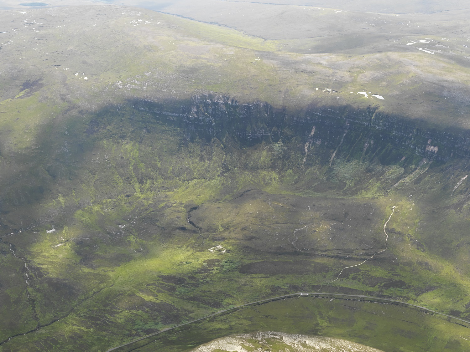 An aerial photograph showing a broad, green moorland landscape with a steep, shadowed cliff face curving across the centre. Faint paths and rocky patches are visible on the slopes below. The location of the Dwarfie Stane lies within this valley landscape.