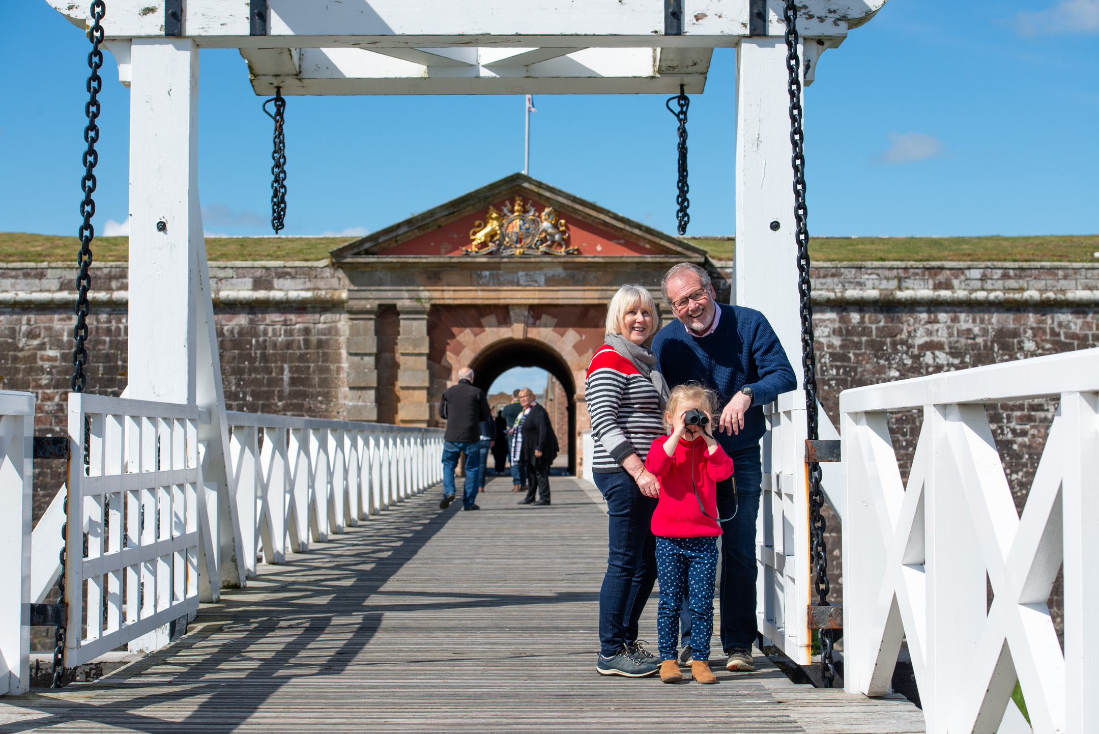 A family pauses on the wooden bridge, with the gatehouse and its colourful crest visible in the background.