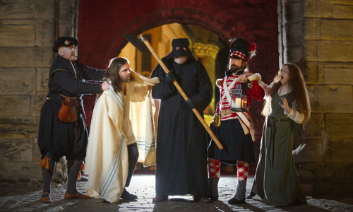 Five costumed performers wearing clothes from across a range of historical eras pose in front of Stirling Castle. The performer in the centre is dressed in a black cloak, black hood and holds an axe. The other four performers look towards them with expressions of shock and fear.
