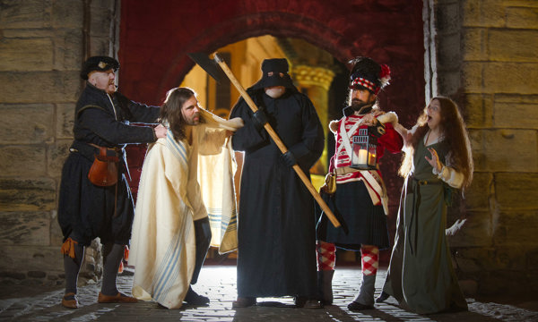 Five costumed performers wearing clothes from across a range of historical eras pose in front of Stirling Castle. The performer in the centre is dressed in a black cloak, black hood and holds an axe. The other four performers look towards them with expressions of shock and fear.