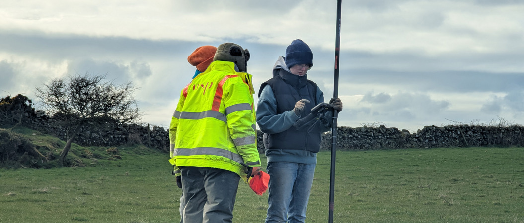 Three people stand in a field looking at an device together, all three are wearing beanies and one wears a high visibility jacket. A small tree in the background has no leaves.