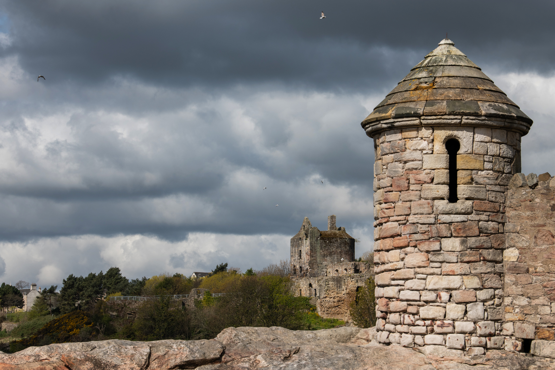 A close-up view of a round stone turret with a narrow vertical arrow-slit built into the wall. The turret stands on a rocky slope with Ravenscraig Castle’s main tower visible in the distance. Overhead, dark storm clouds gather while seabirds circle in the sky.