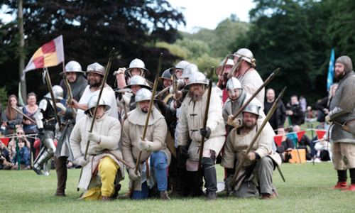 A group in costume on a battlefield raise their spears to the camera 