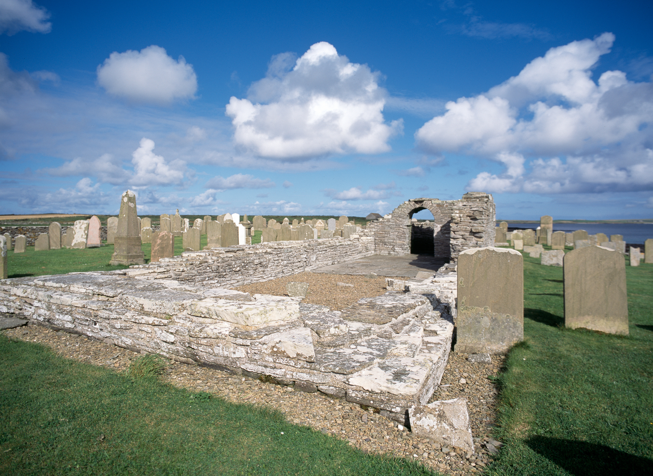 Close view of the low stone walls of Westside Church’s rectangular structure, surrounded by upright gravestones on a green lawn beneath a blue sky with scattered clouds.