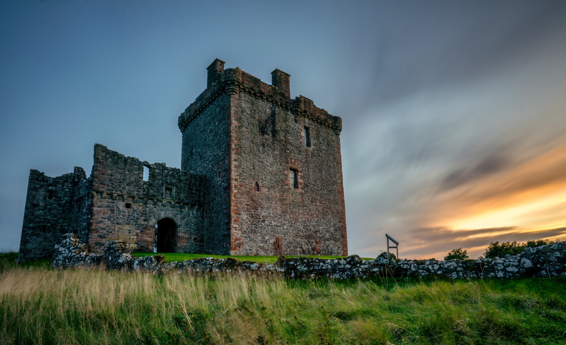 A dramatic sunset sky over an imposing tower house. The main house remains strong and imposing but other parts are more crumbling and ruinous. 