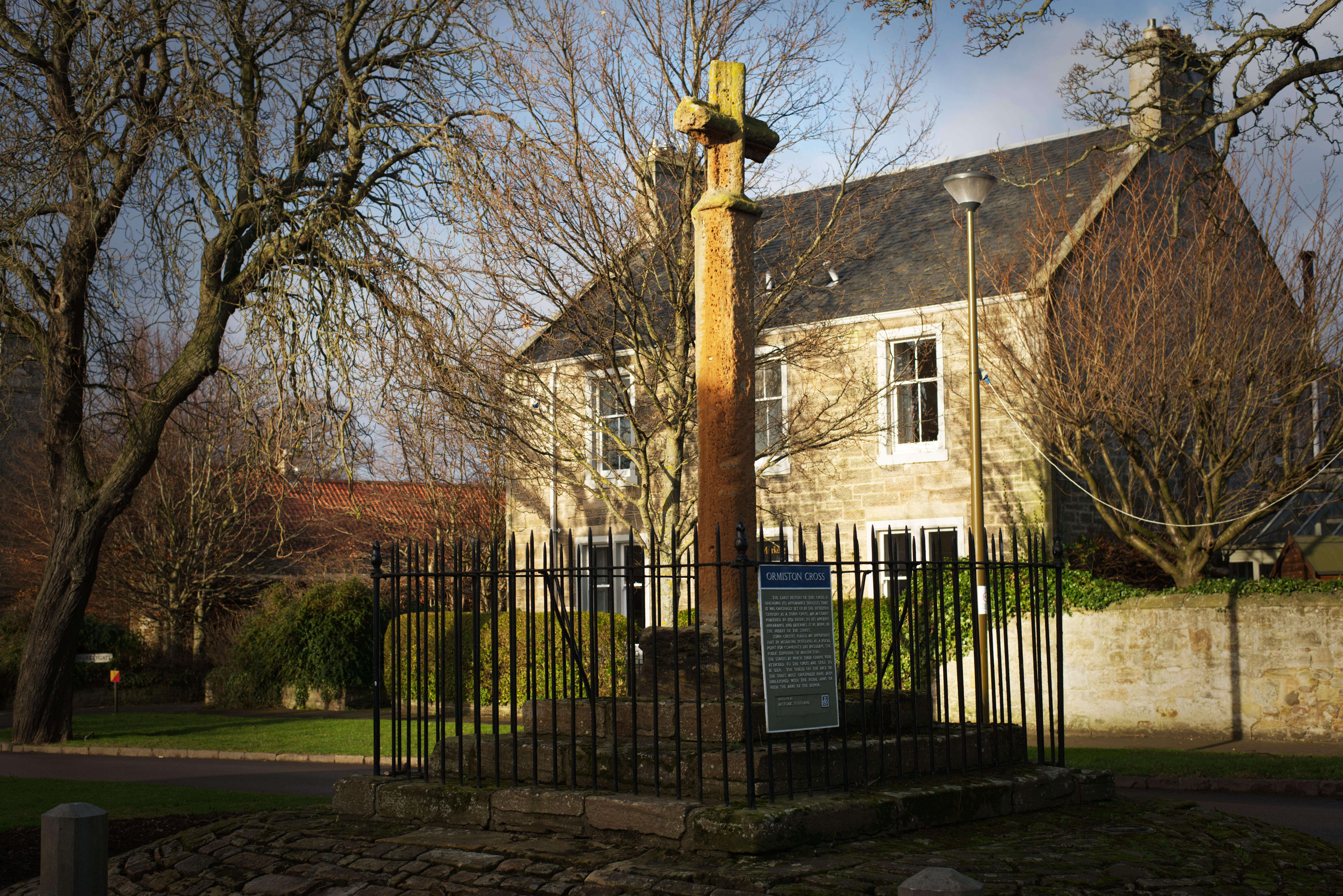 Stone market cross standing on a raised circular base and enclosed by a black iron fence in a village setting. The tall weathered shaft is topped with a small carved cross. Behind it is a two‑storey stone building with white‑framed windows, leafless trees, and a streetlamp. Low afternoon sunlight casts warm tones across the scene, highlighting the cross and surrounding greenery.