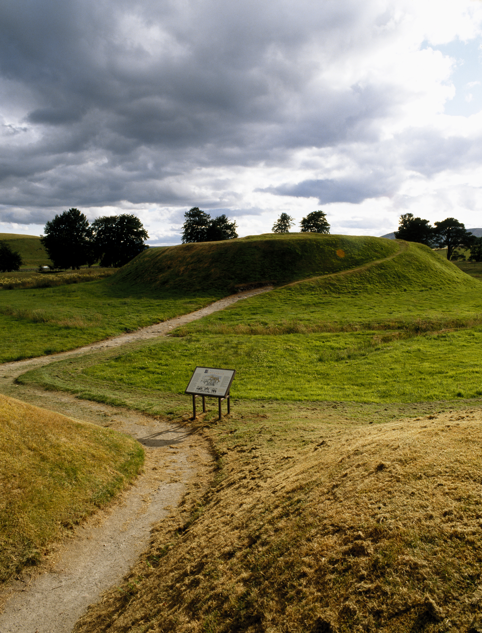 Lumphanan earthwork mound with a winding dirt path leading toward it, an information board in the foreground, and trees in the background under a cloudy sky.