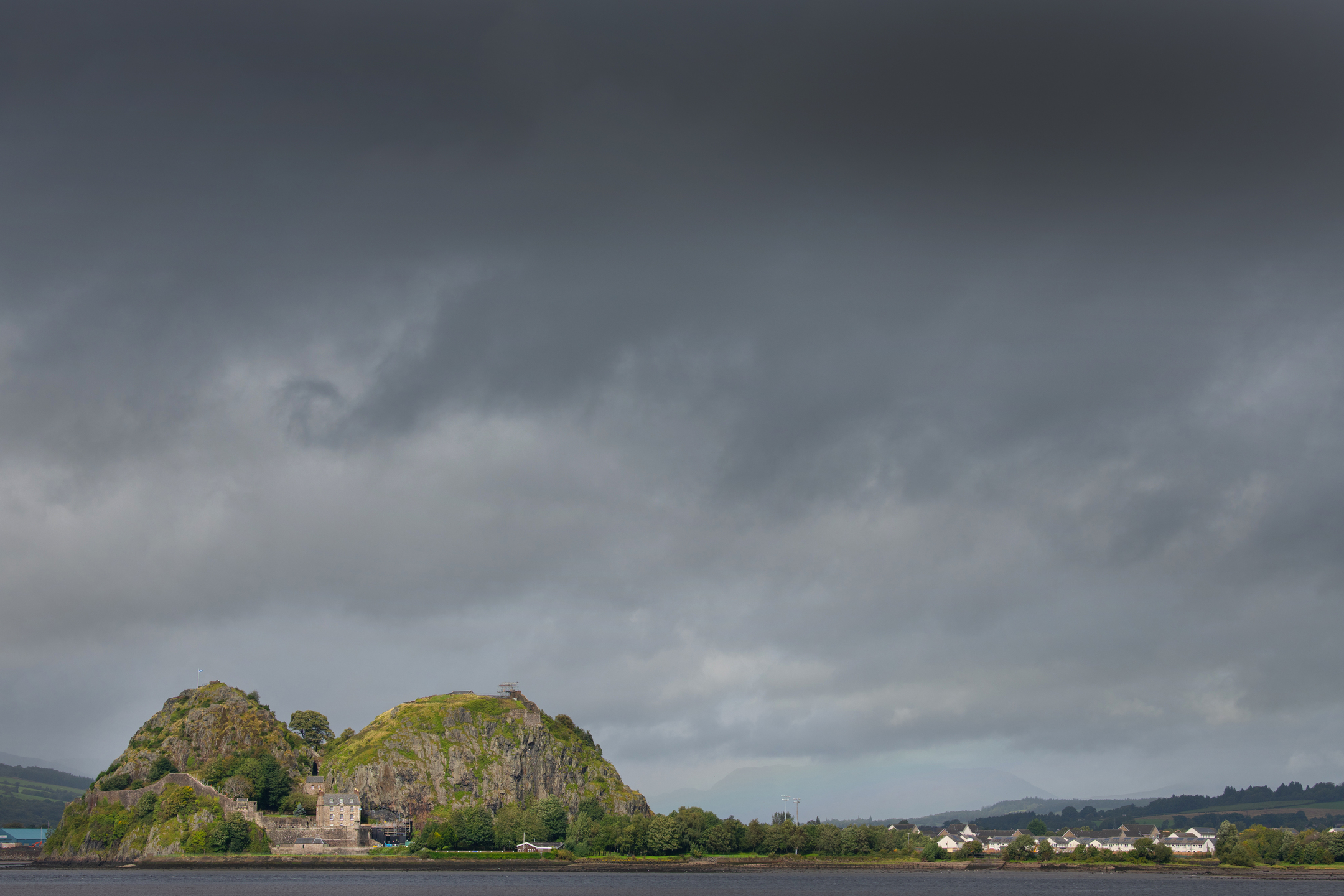 A dramatic, cloudy sky hangs over Dumbarton Castle, which sits on twin rocky outcrops beside the water.