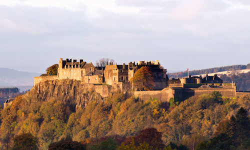 A stone castle stands atop a hill. Below is a forest of trees and behind are rolling hills. The sky above is blue and the sun is shining.