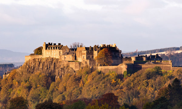 A stone castle stands atop a hill. Below is a forest of trees and behind are rolling hills. The sky above is blue and the sun is shining.