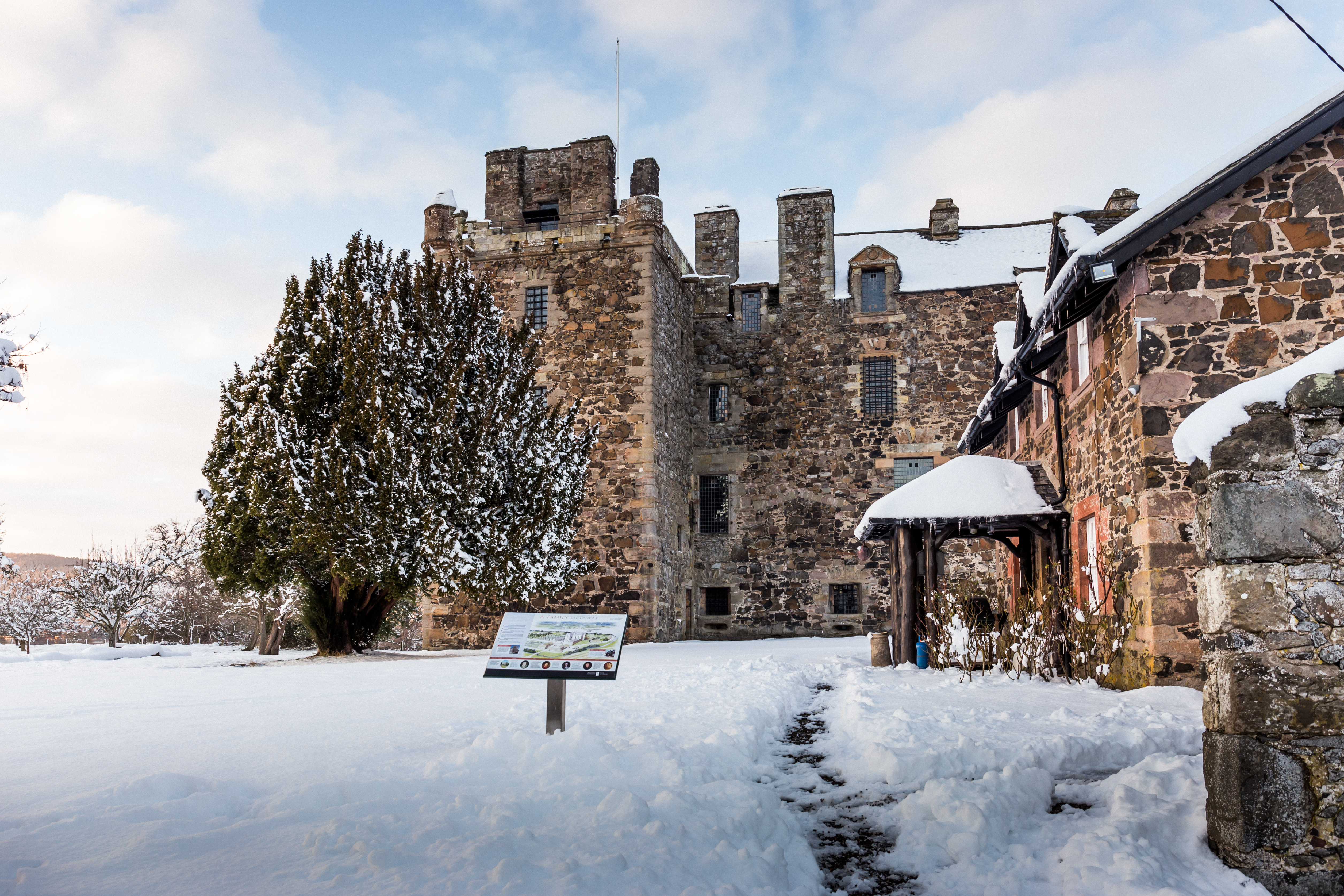 Front view of Elcho Castle and its adjoining buildings covered in snow, with a cleared path leading to the entrance. An interpretation panel stands in the foreground under a bright blue sky with scattered clouds.