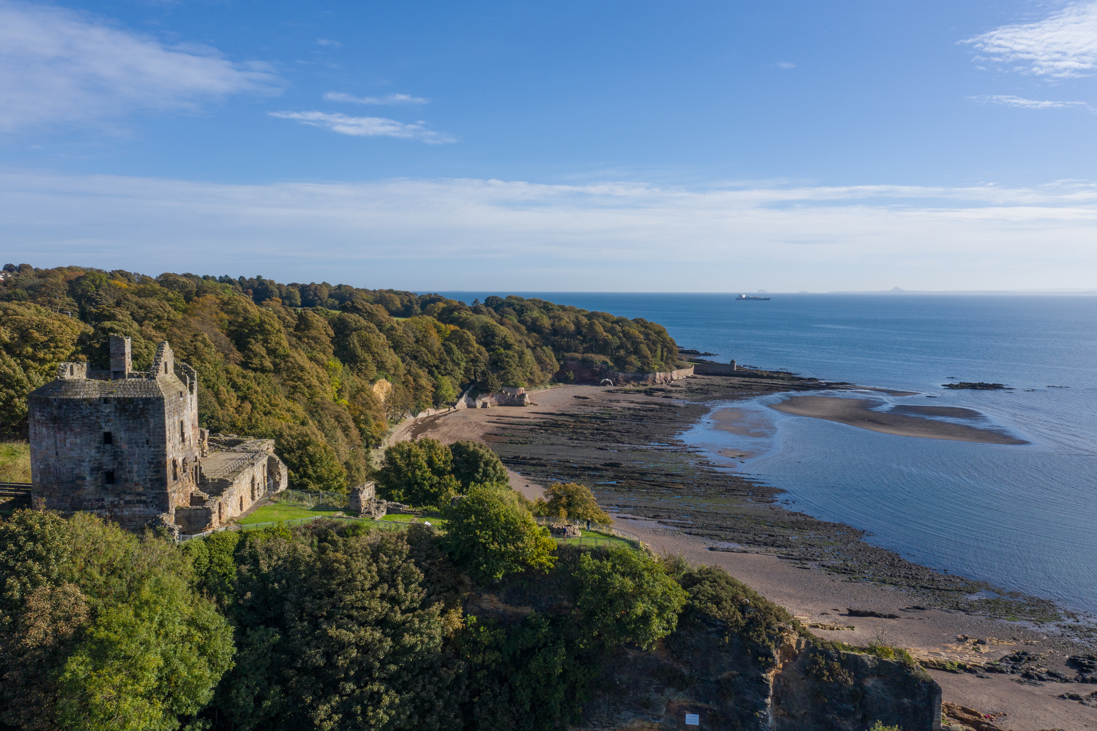 An elevated coastal view of Ravenscraig Castle’s ruined stone tower on a wooded headland overlooking a wide bay. The shoreline curves into the distance with exposed sand and dark rock beds at low tide. Dense autumn-coloured trees cover the hill behind the ruins, and the calm blue sea stretches to the horizon under a bright sky.