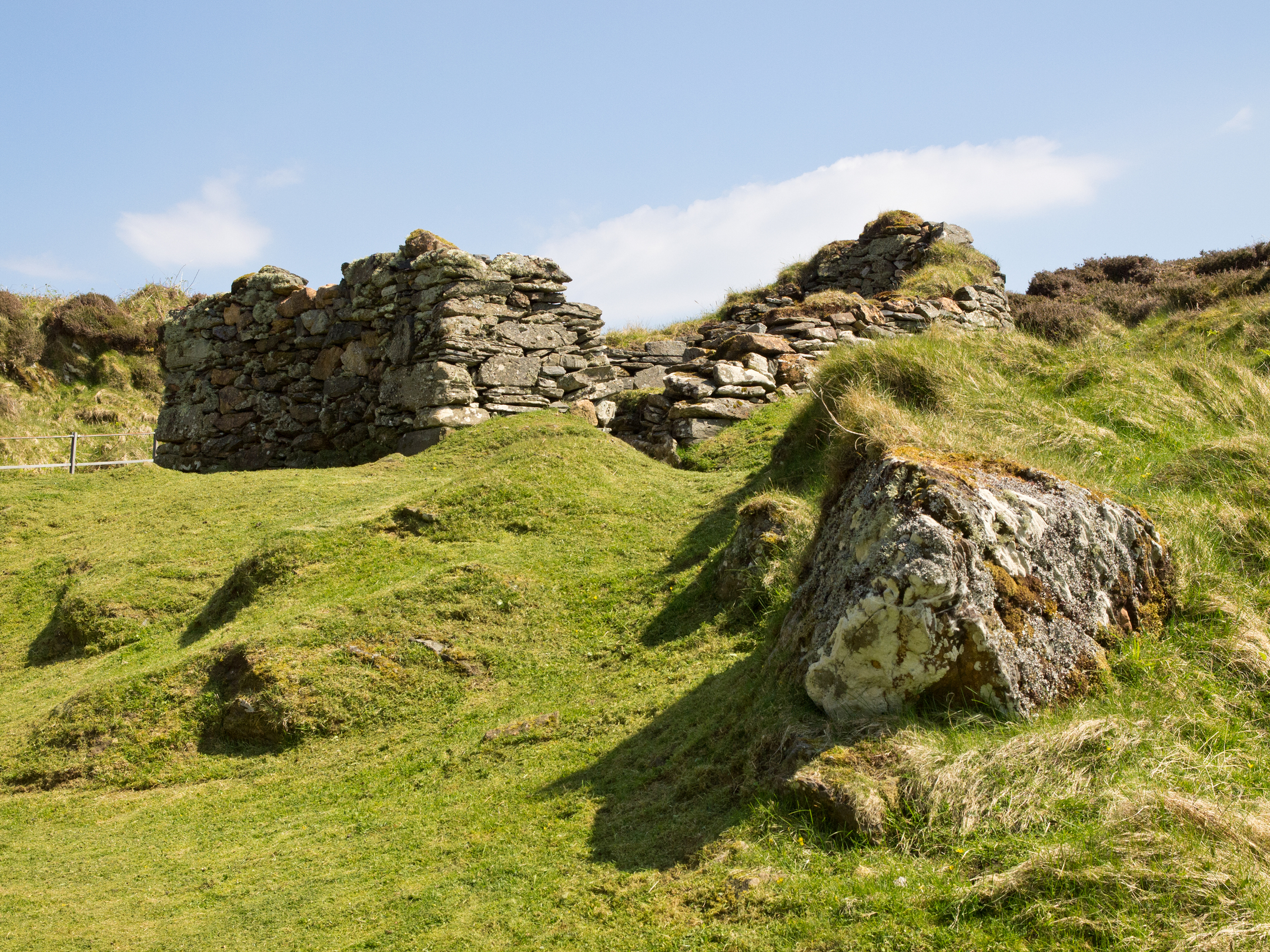 Partially collapsed stone walls of an ancient structure on grassy slopes with rocky terrain in the background.