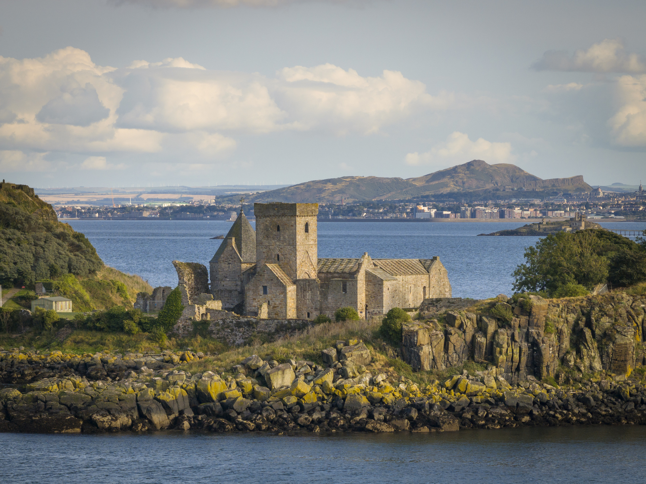 A large, stone historic abbey on a small island. Across the water, in the distance, there are buildings and hills