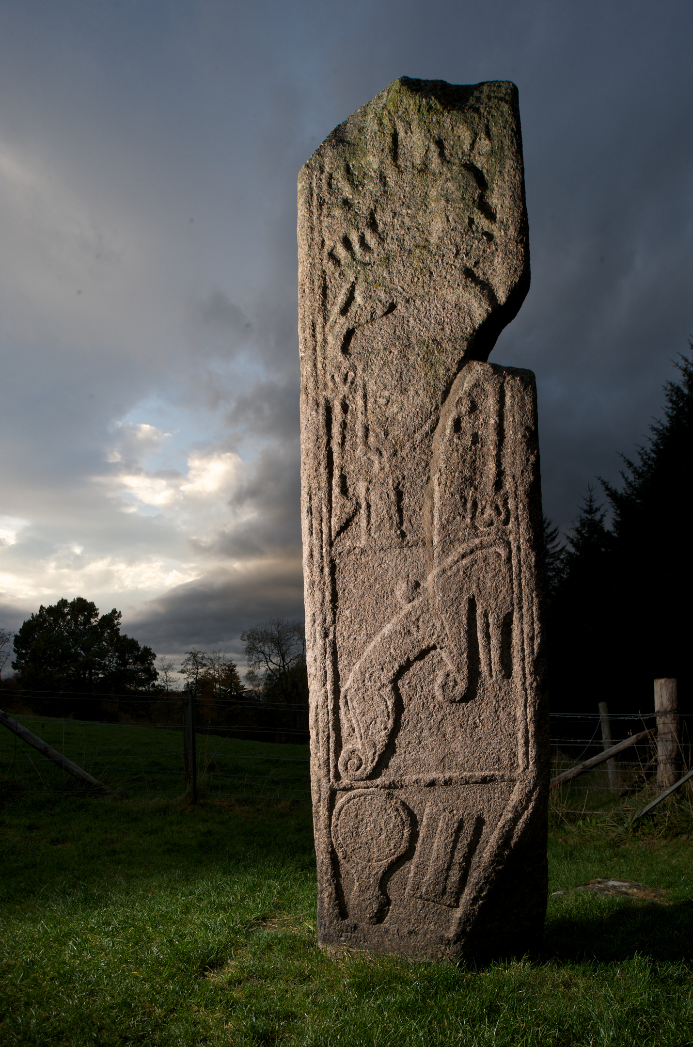 The Maiden Stone standing upright in a field at dusk, illuminated from the side, showing its carved Pictish symbols