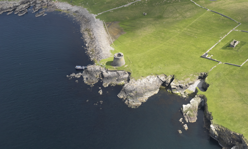 An aerial photograph of a round stone fort stood on a cliff side. To the right and above are green fields with some stone walls running through them. To the left and below are cliff edges and beaches which meet the sea.