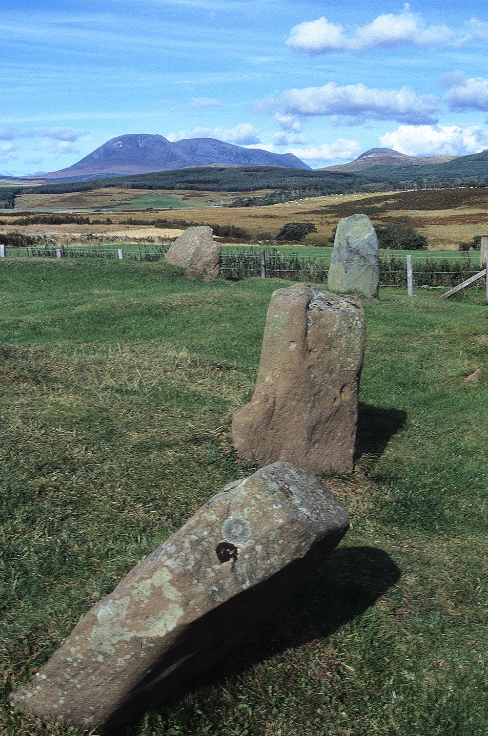 Several upright stones in a grassy field with rolling hills and mountains in the background beneath a blue sky.