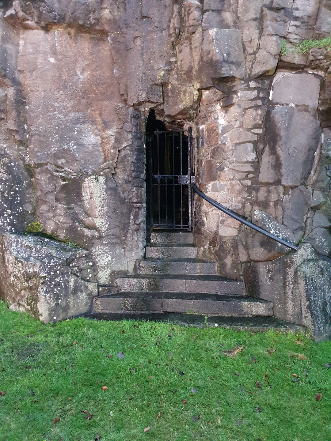 There is a door with a bar door in front of it, in the middle of an old stone wall. There are stairs leading up to it, with grass in front of it.