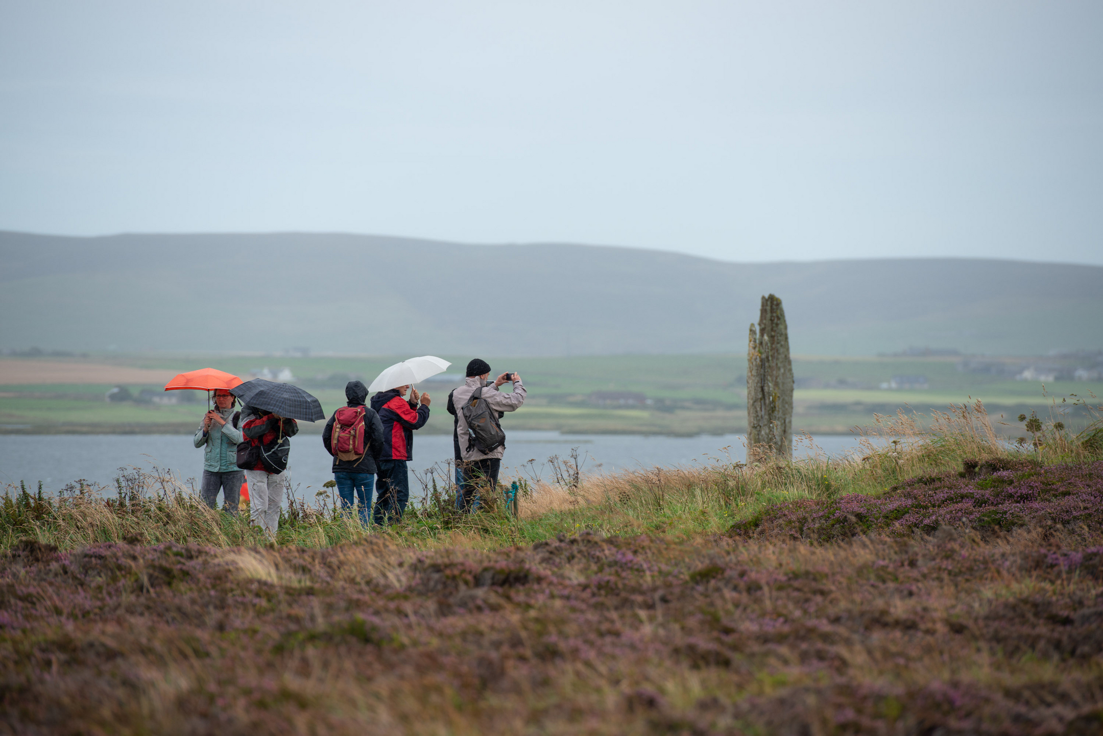 A group of visitors photograph a stone circle on a rainy, yet atmospheric day. A mix of umbrellas and anoraks helps keep the worst of the weather out. 
