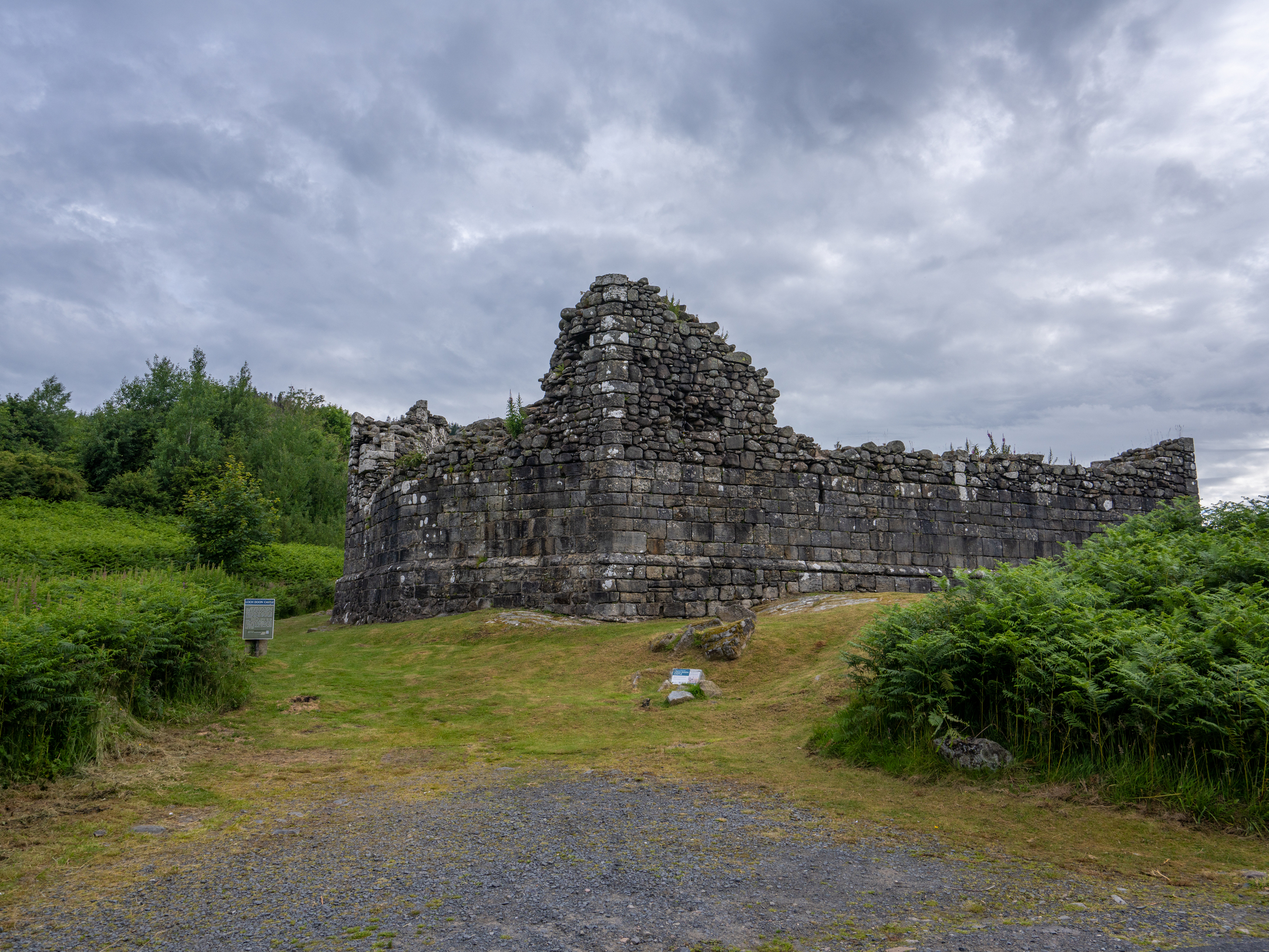 A side view of the stone walls of Loch Doon Castle, showing the ruins surrounded by grass and bracken.