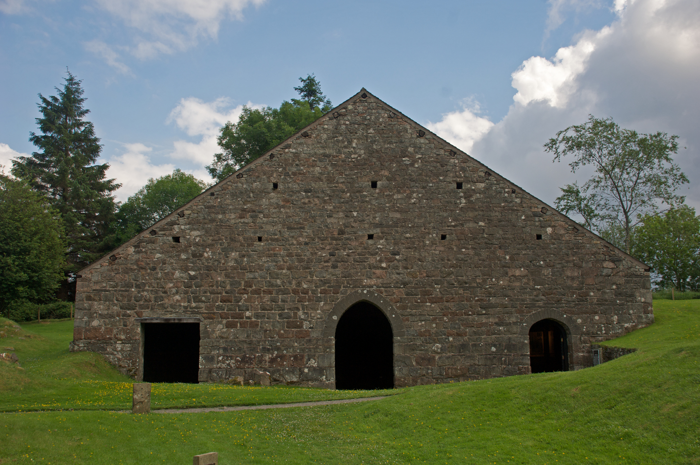 A large, triangular‑fronted stone building with three dark arched doorways along the base. The wall is made of rough, uneven stones with small square openings scattered across it. The building sits on a grassy slope, surrounded by trees