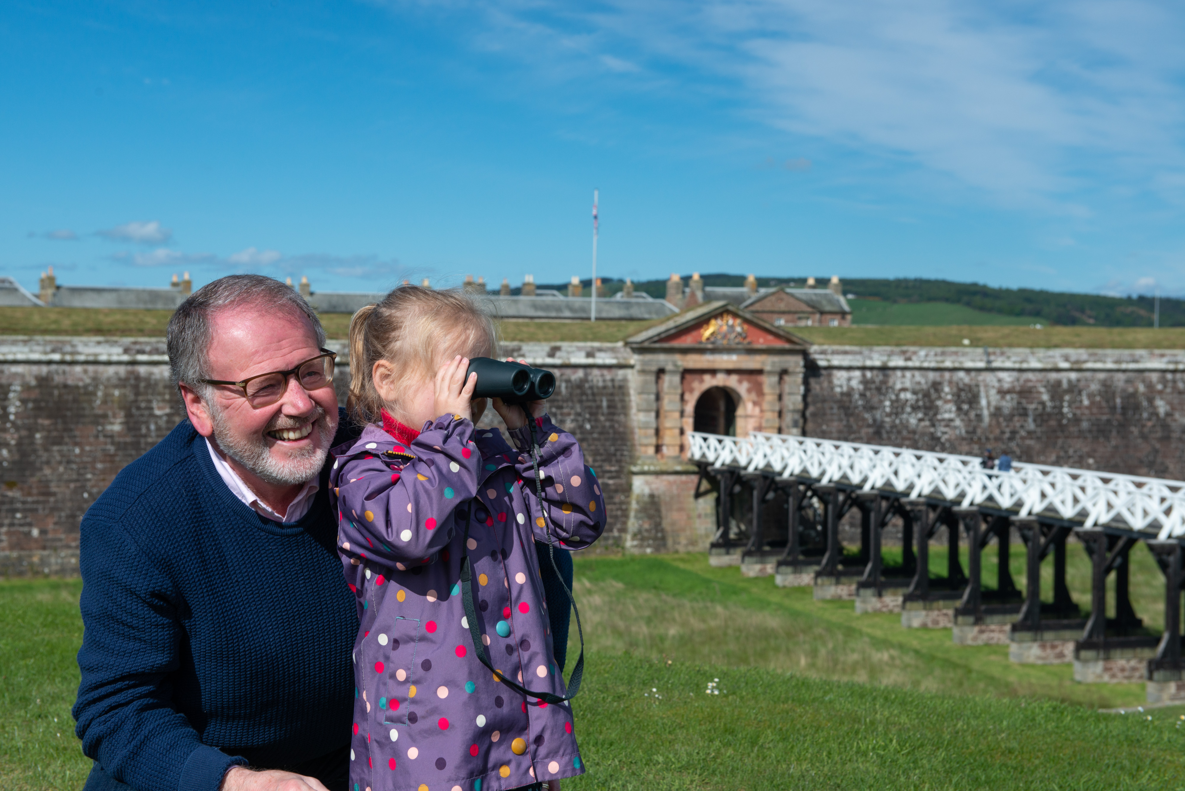 A smiling adult crouches beside a small child using binoculars on a grassy fortification, with a drawbridge and stone gateway behind them.