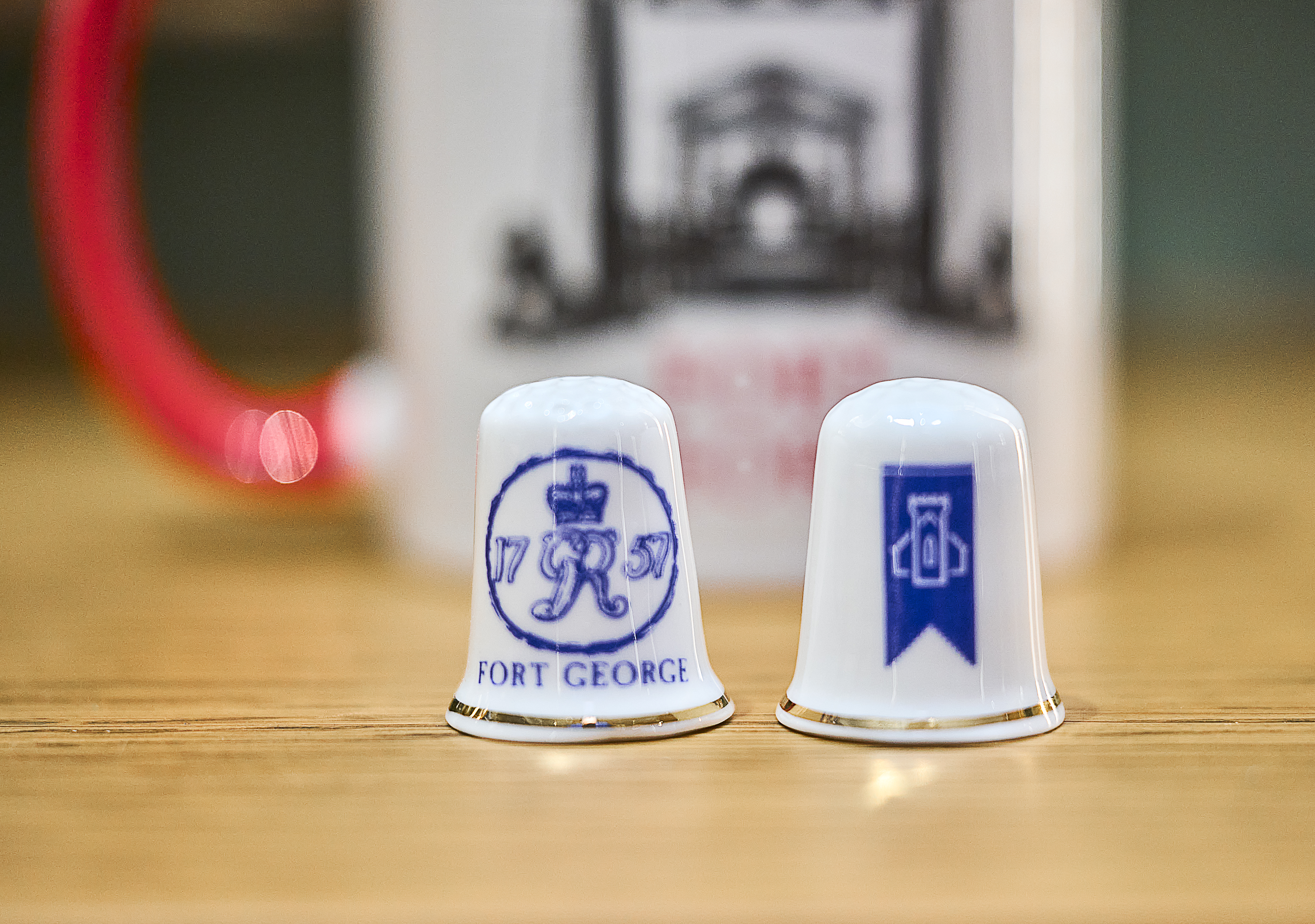 Two ceramic thimbles on a wooden surface. One features the Fort George crest, the other displays a blue emblem.