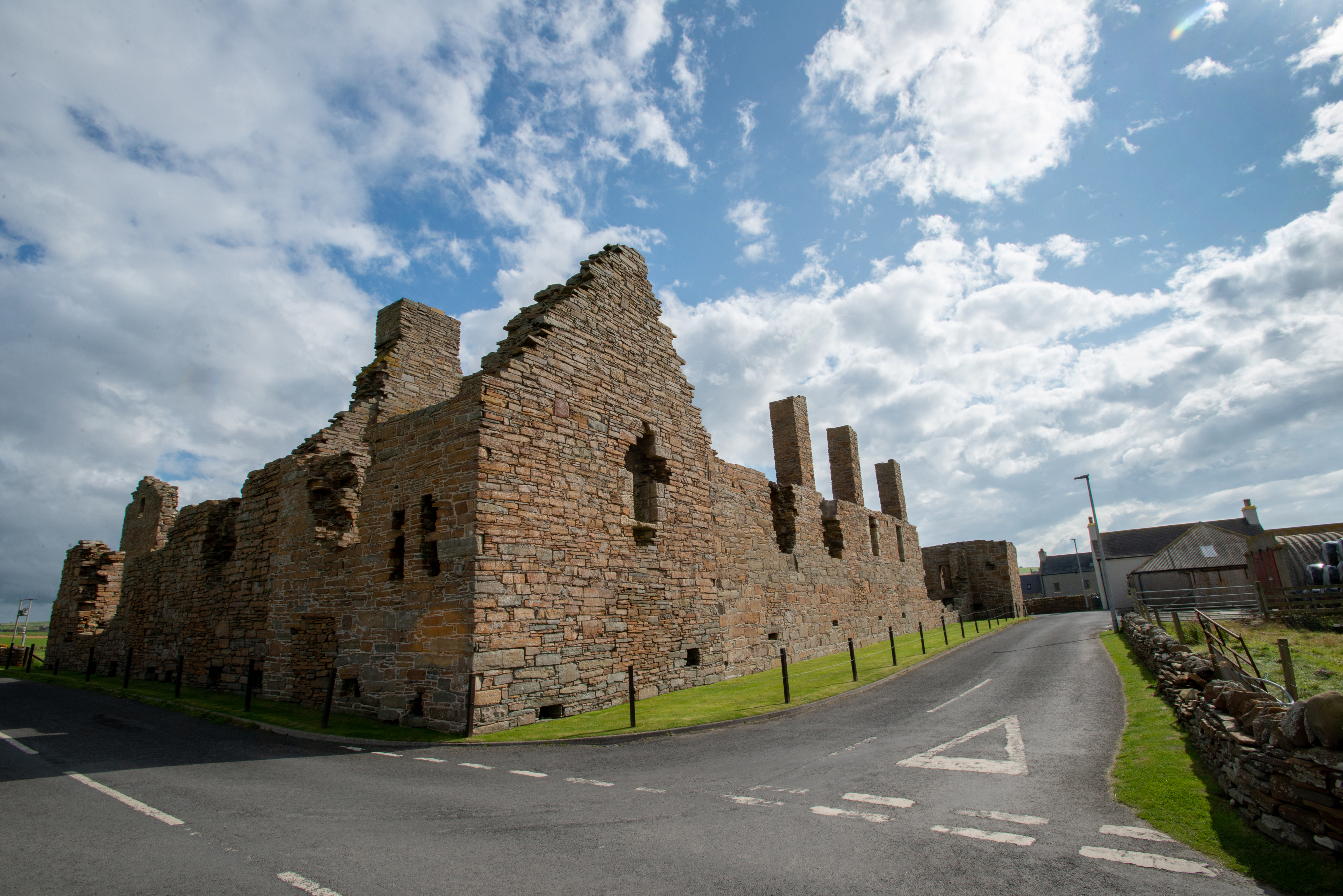 A roadside perspective of Earl’s Palace, with the long outer wall standing against a dramatic sky of clouds and blue patches. The building’s jagged top and vertical stone remnants rise above the road below.