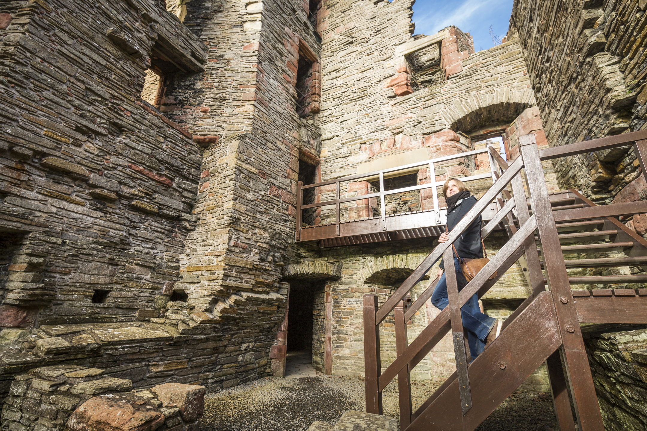 A person standing on a wooden staircase inside a tall stone ruin, surrounded by weathered brick walls and open archways.