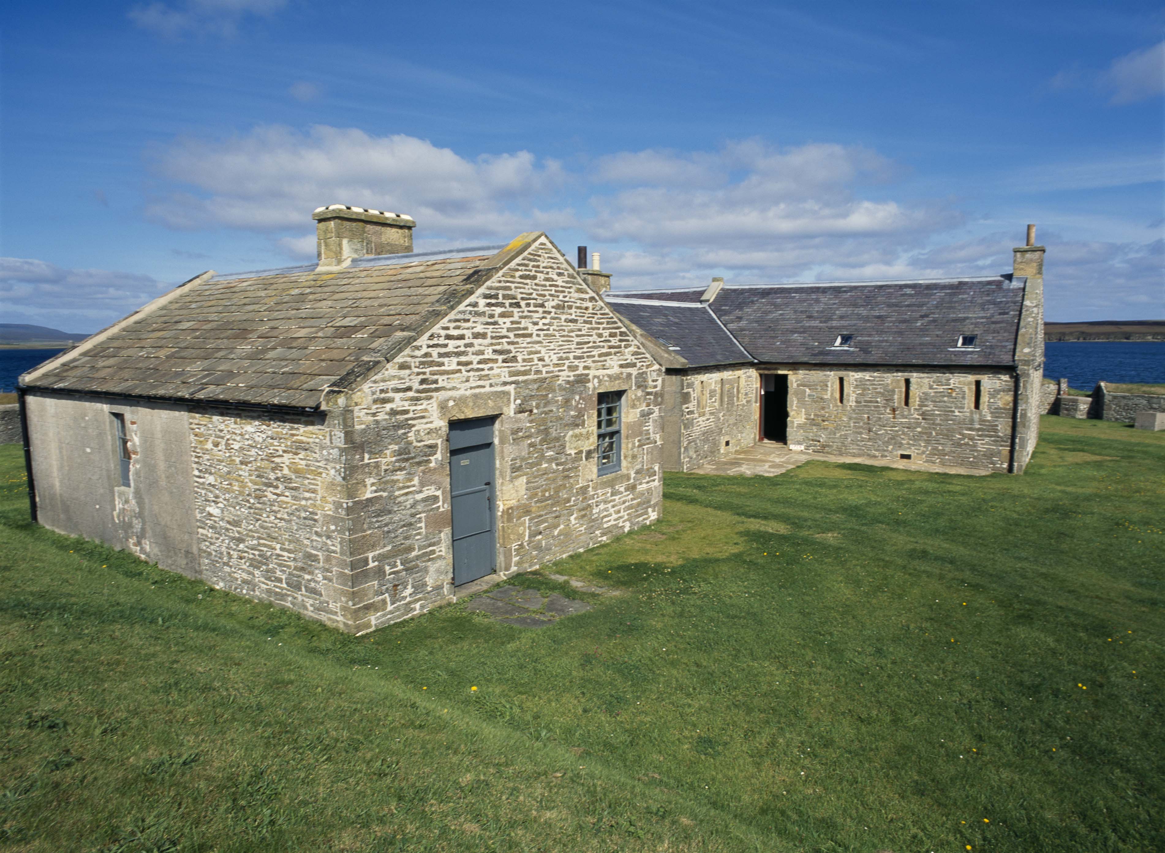 A group of single storey stone buildings with slate roofs, on a grassy area, by the sea