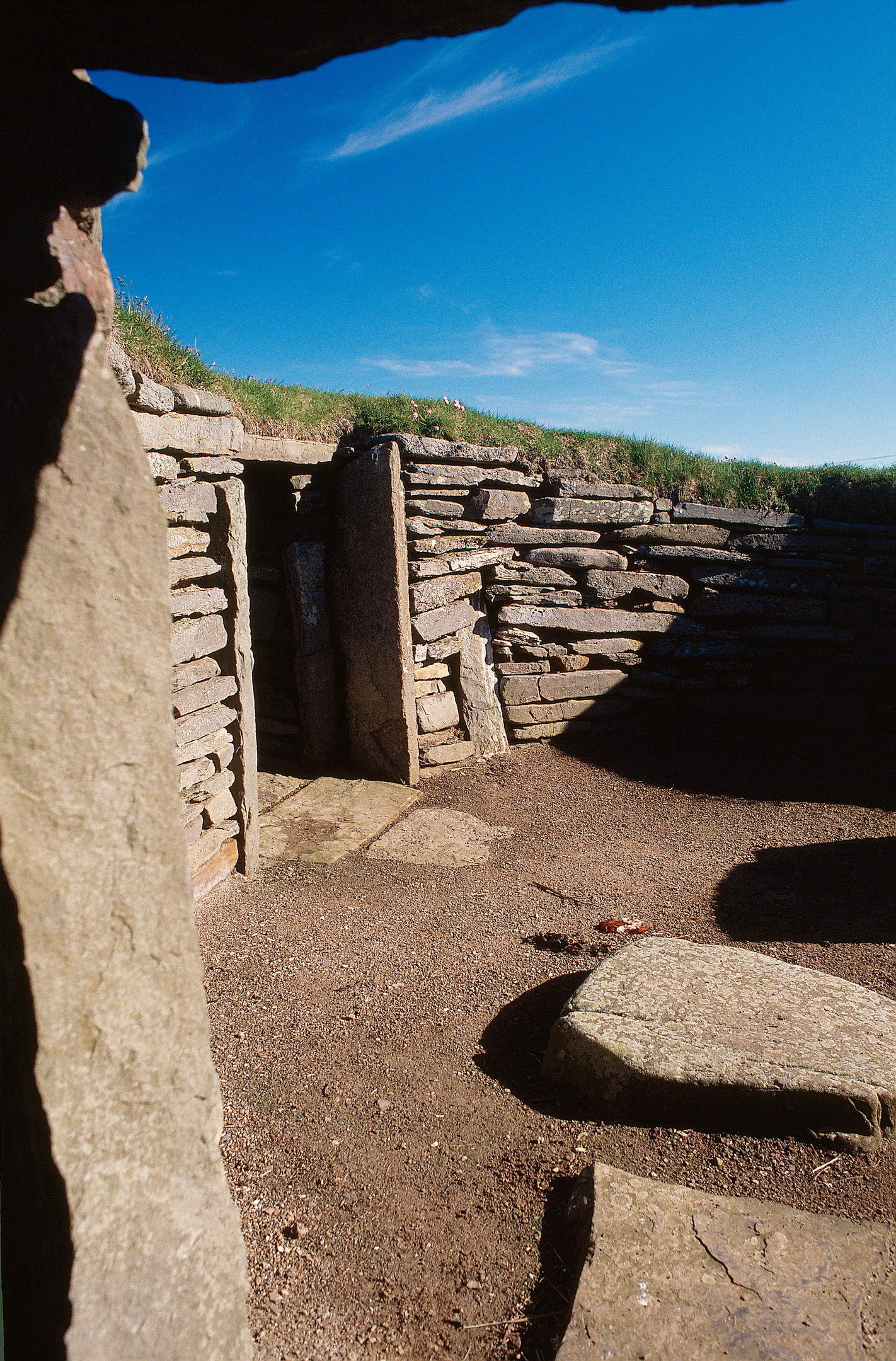 Interior view of the Neolithic stone house, showing stacked stone walls, upright doorposts, and sunlight streaming in from above.