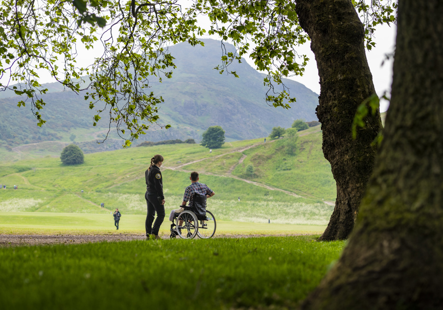The photograph shows two individuals beside a tree looking over Holyrood park