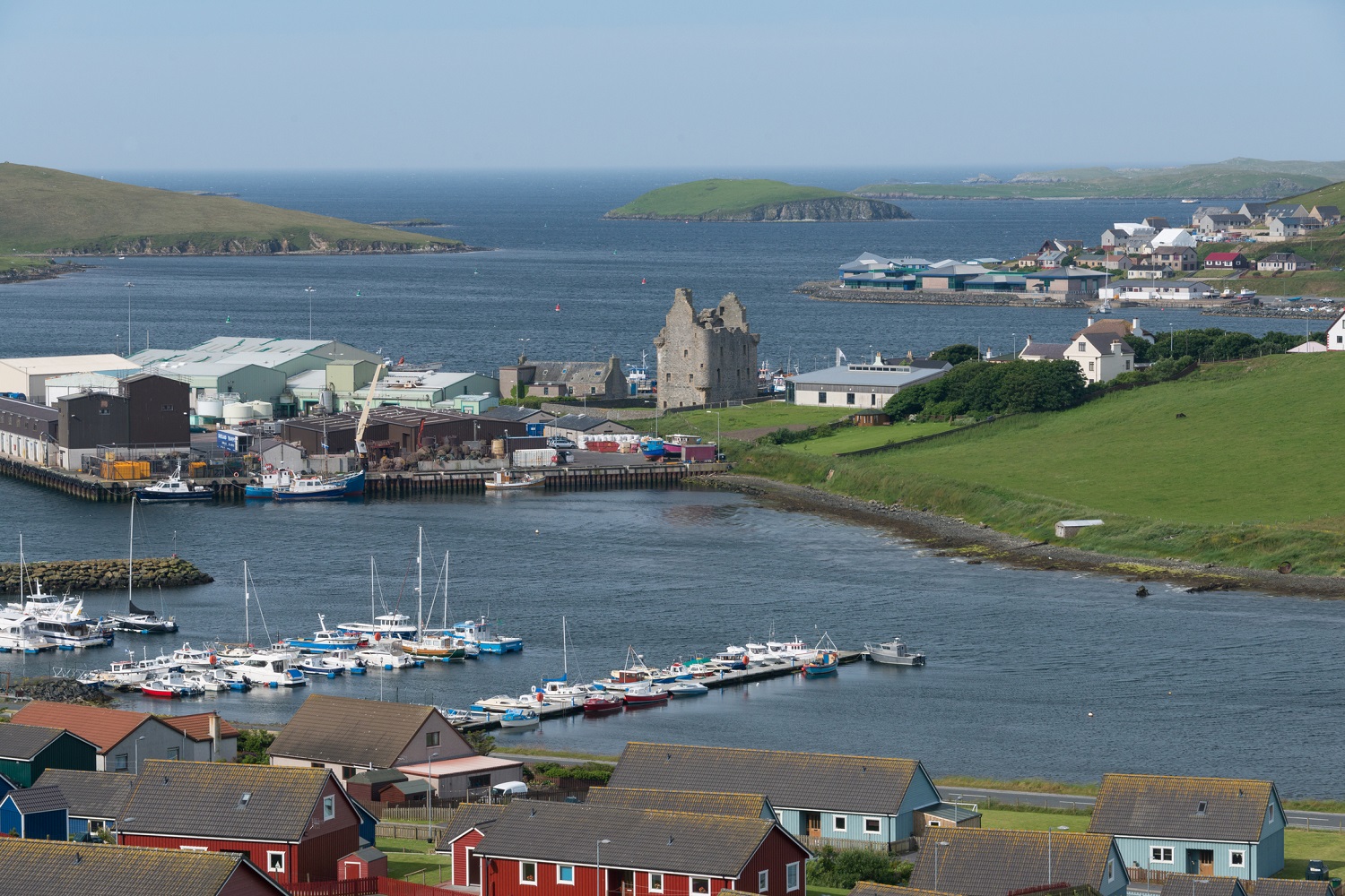 Scalloway Castle overlooking a marina and colourful houses, with green hills and small islands in the background.