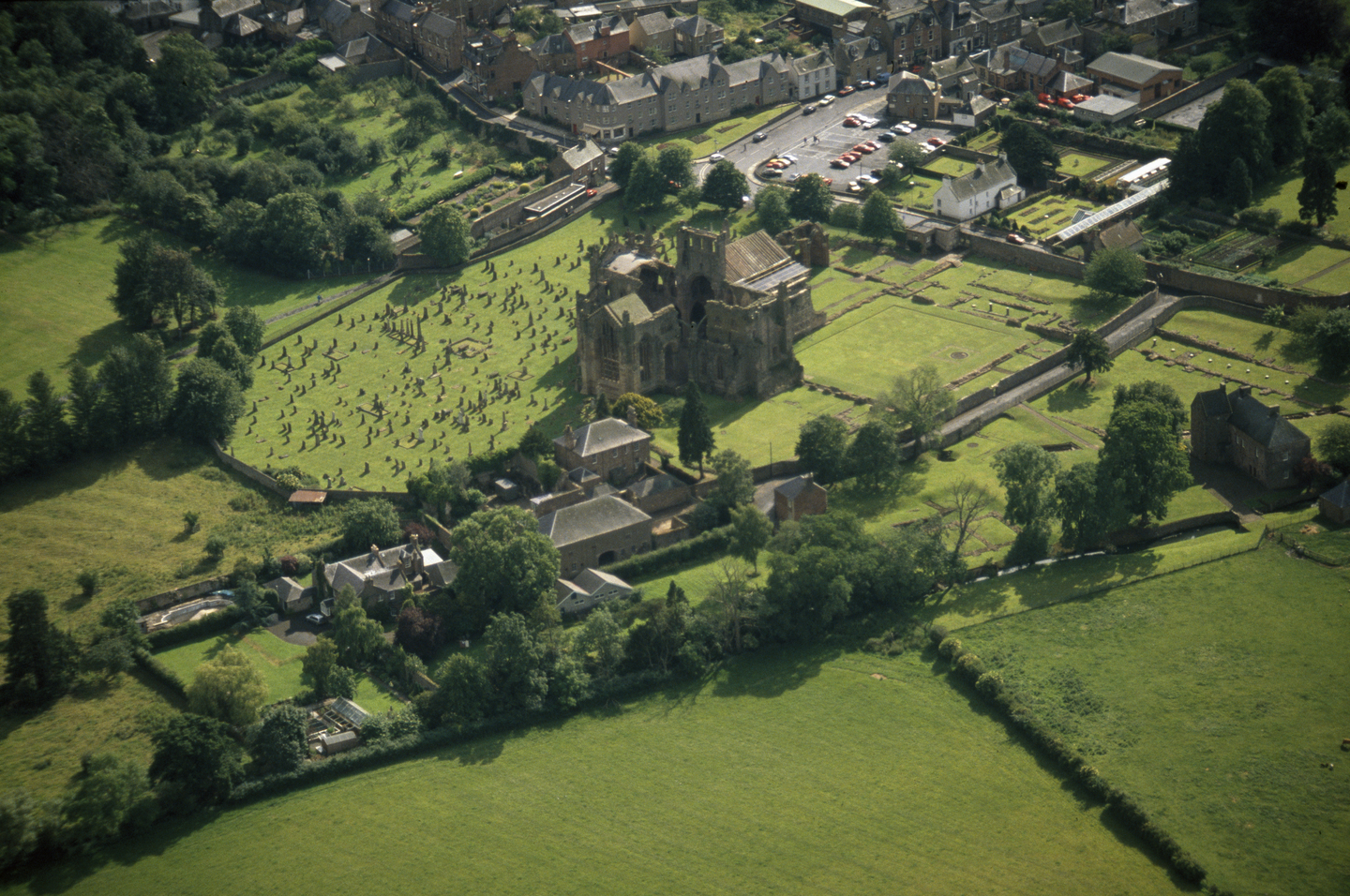 Aerial view of a large stone historical building which resembles a church or abbey. There is a graveyard next to it, as well as green fields with trees surround them more widely. In the top right there is also a car park with housing. 