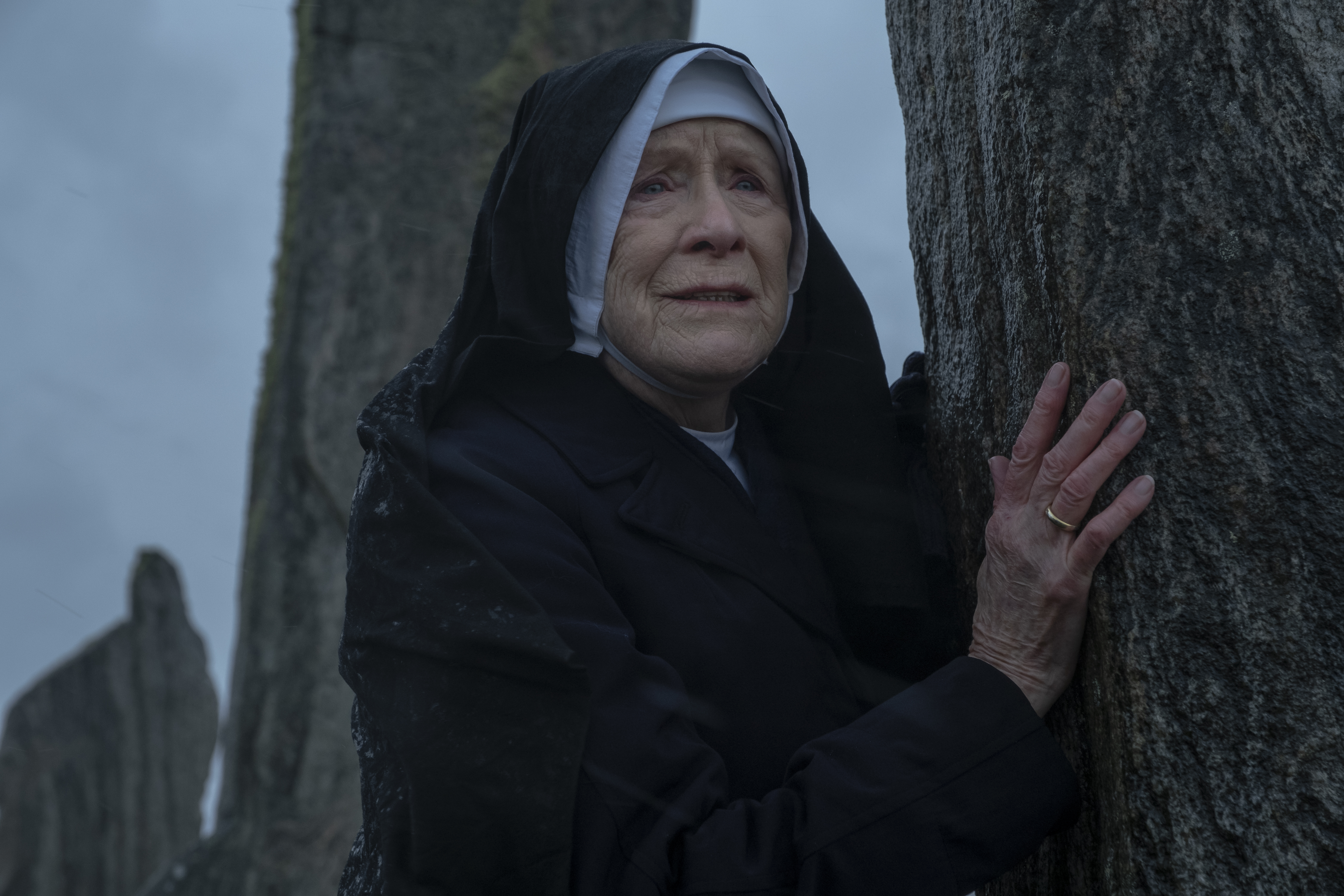 An elderly nun clutches a tall stone pillar outdoors, looking emotional as wind and rain sweep across a bleak, rocky landscape.